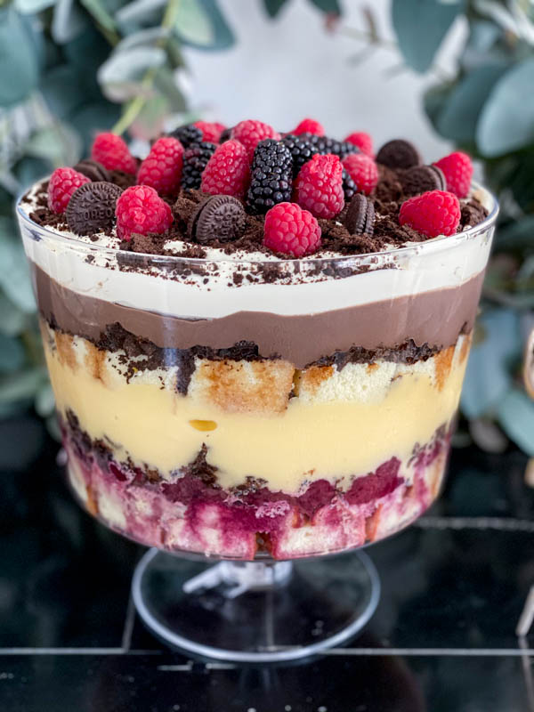 A close up of the trifle showing all the layers and topping. It is topped with fresh raspberries and blackberries with crushed Oreo biscuits and mini Oreo biscuits scattered around.