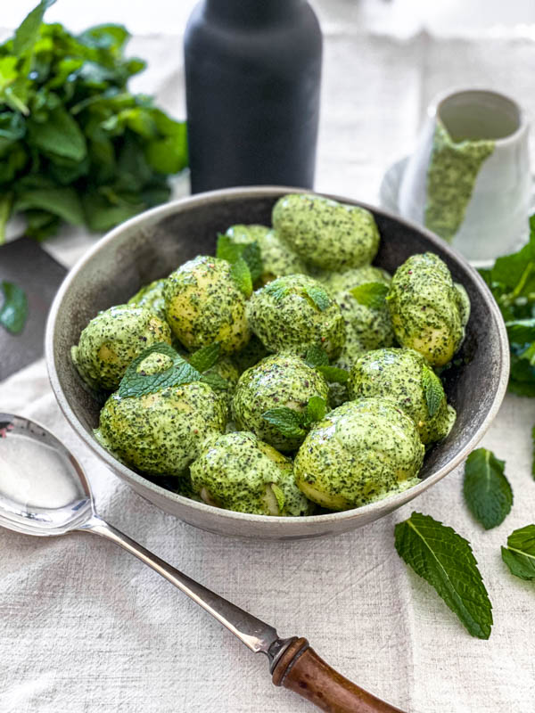 A bowl of boiled baby potatoes that have been dressed with the Easy Mint Dressing. In the background are fresh mint and a jug with the dressing inside.