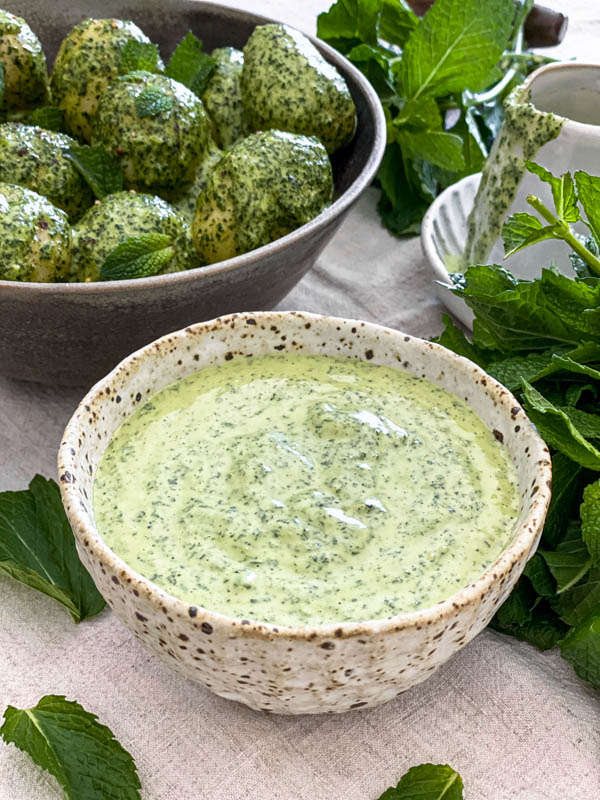 The Mint Dressing is in a bowl with mint leaves scattered around it and a bowl of the Potato Salad in the background.