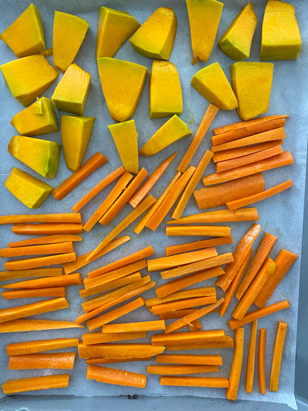 Carrot sticks and pumpkin pieces are evenly spread out on an oven tray lined with baking paper, ready for cooking in the oven.