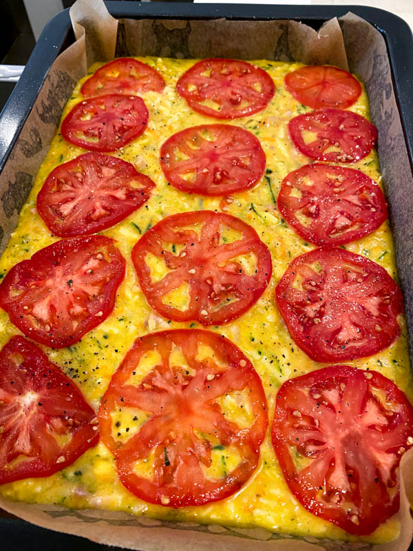 The zucchini slice batter is now in a rectangle baking pan and is topped with sliced tomatoes, ready to be cooked in the oven.