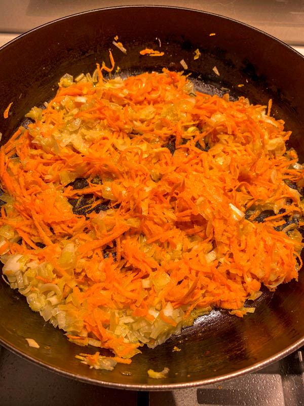 The chopped onions and grated carrot are being sautéed in a frying pan.