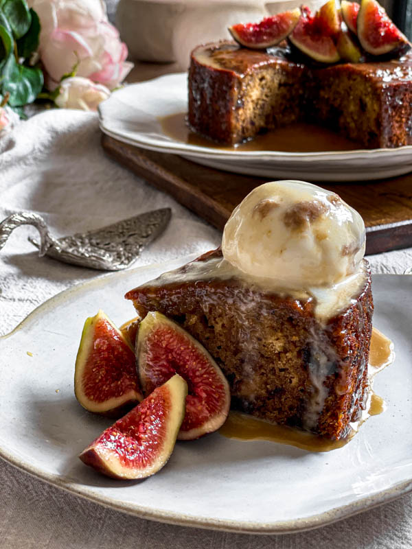A slice of pudding is on a serving plate with a scoop of ice-cream on top of it and fresh figs alongside. In the background is the pudding, a cake spatula and flowers.