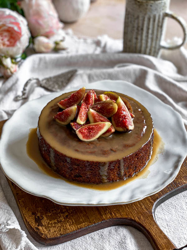 The decorated pudding is on a plate that is sitting on a wooden board. There are flowers, a cake spatula and jug in the background.