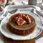 The Sticky Date Pudding is on a plate on top of a wooden board and sitting on a linen tablecloth.