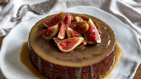The Sticky Date Pudding is on a plate on top of a wooden board and sitting on a linen tablecloth.
