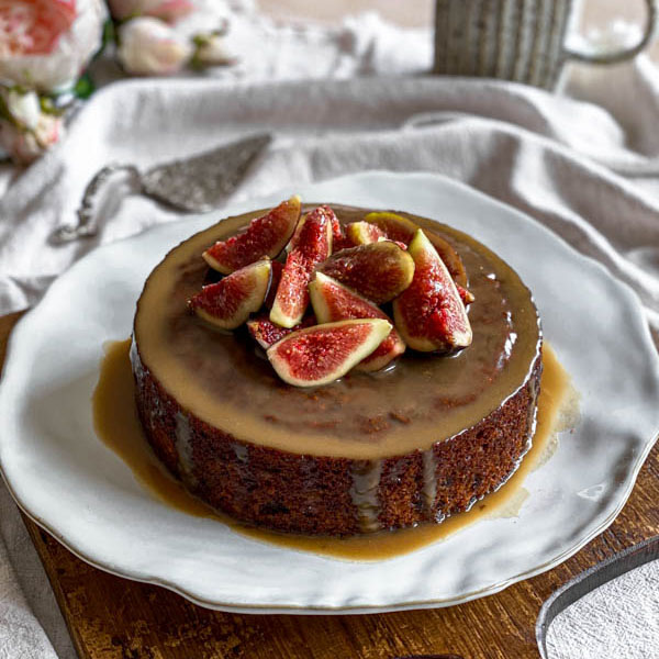 The Sticky Date Pudding is on a plate on top of a wooden board and sitting on a linen tablecloth.