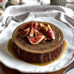 The Sticky Date Pudding is on a table with a cake spatula, ceramic jug and flowers in the background.