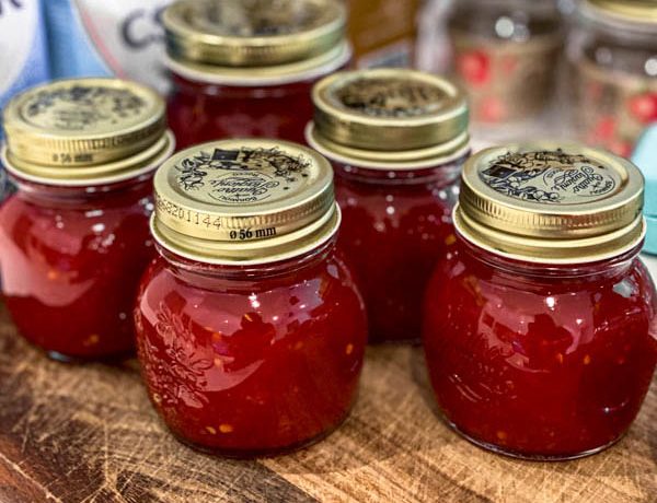 The bottled jars of Tomato Chilli Jam are sitting on top of a wooden board.