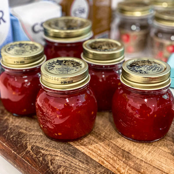 The bottled jars of Tomato Chilli Jam are sitting on top of a wooden board.