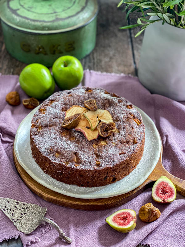 The cake is on a serving plate sitting on top of a round wooden board. The cake has been dusted with icing sugar and decorated with dried apple slices and semi-dried figs.