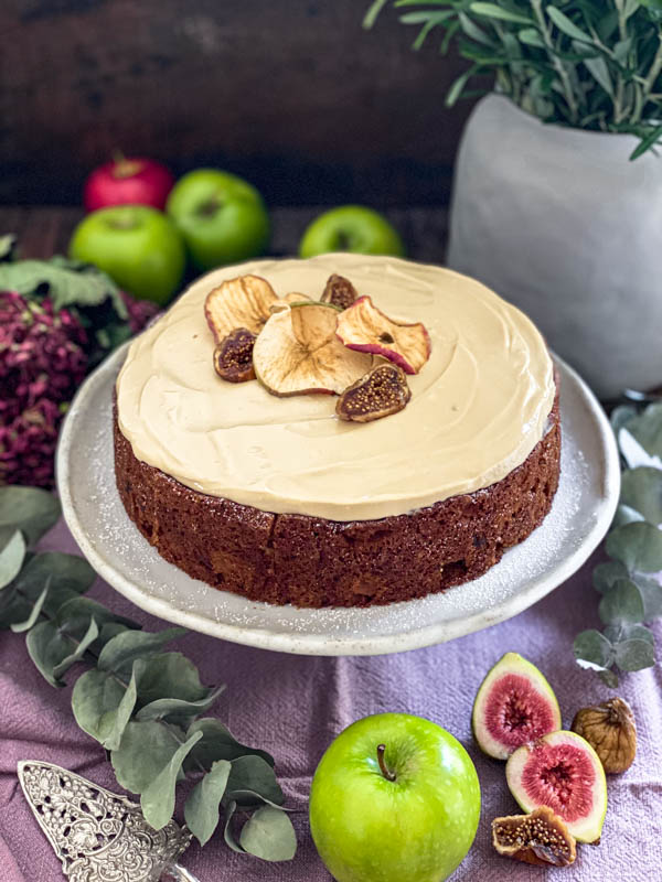 The apple cake has been topped with Maple Cream Cheese frosting and is on a cake stand. It is decorated with dried apple slices and sliced semi-dried figs which are sitting in the middle of the frosting.