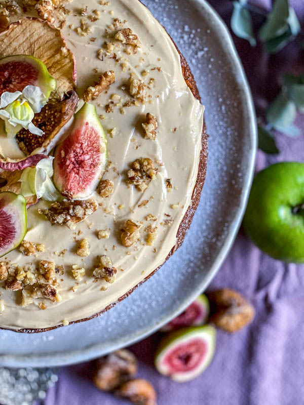 Looking down onto the top of the cake. It is a close up of half of the decorated cake. The frosting has been sprinkled with chopped walnuts and decorated with fresh fig quarters, semi-dried figs, dried apple slices and small white flowers.