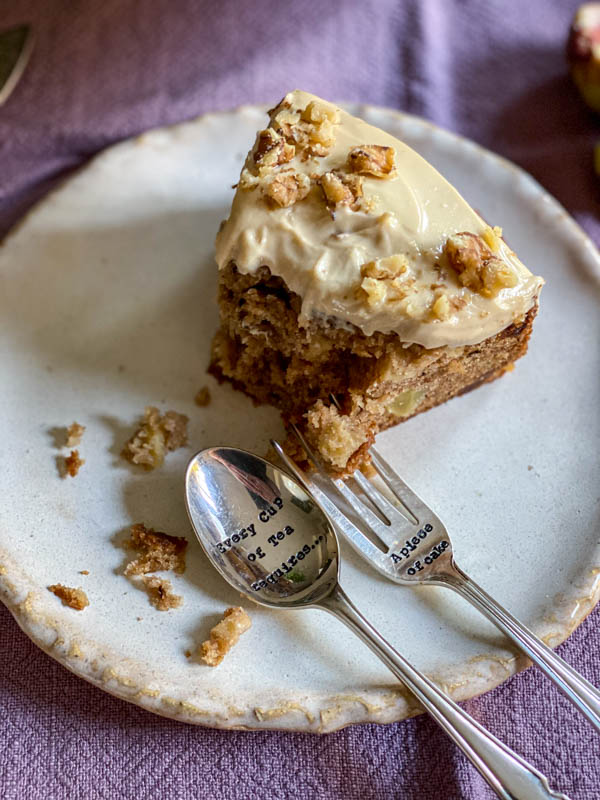 Looking down onto a half eaten slice of apple cake on a serving plate with a cake fork and spoon resting on the plate.