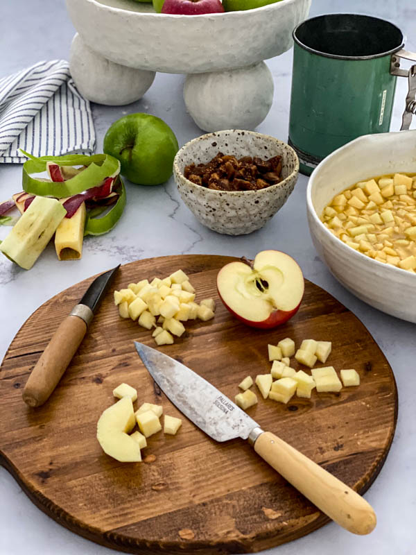 The apples are being diced on a wooden chopping board. In the background are a bowl of the chopped apples, a bowl of chopped figs along with apple peelings and cores.