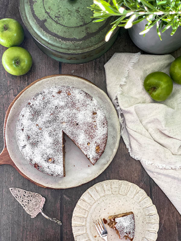 Looking down onto the apple cake that has had a slice removed. There is also a piece of eaten cake on a small plate, a vintage cake server, green apples, a napkin and cake tin on the wooden table as well.