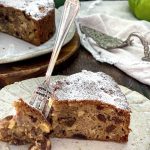 A slice of apple cake on a plate with a silver cake fork on the plate as well.