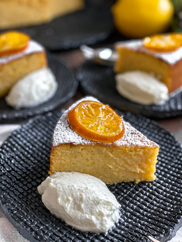 A close up of a slice of Whole Meyer Lemon Cake that has been dusted with icing sugar and also has a slice of confit lemon sitting on top of it. It is served with a dollop of Greek Yoghurt.