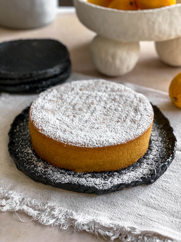 The Whole Meyer Lemon Cake is liberally dusted with icing sugar and is on a black ceramic serving plate on a table with a piece of cloth underneath the plate.