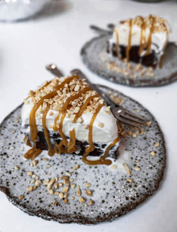 Slices of brownie cake are on plates ready to be served. They have been served with chopped roasted peanuts on top and a drizzle of salted caramel.