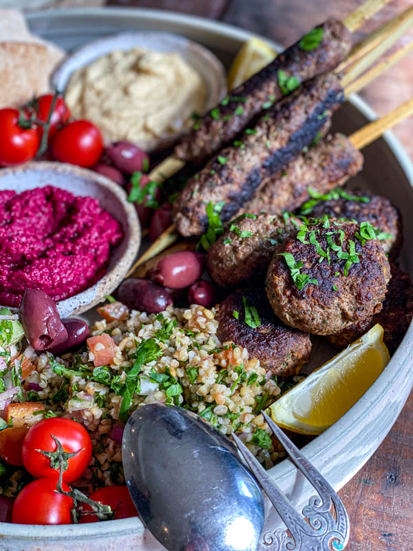 A close-up of the kofta skewers and patties that form part of a Middle Eastern style platter. There is also tabbouleh, olives, tomatoes and dips on the platter.