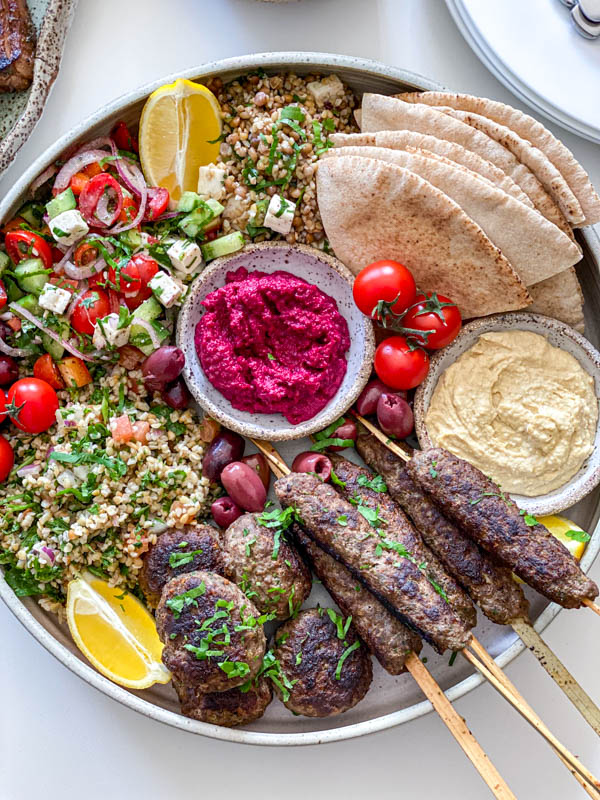 Looking down onto the Middle Eastern inspired platter featuring kofta skewers and patties. There is also pita bread triangles, hummus, beetroot dip, tabbouleh, greek salad, olives and tomatoes on the platter as well.