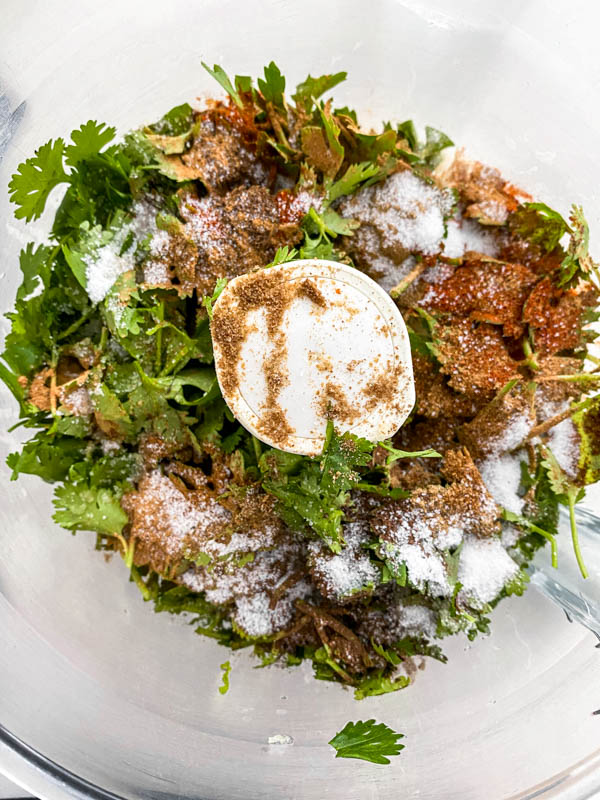 The spices, seasoning and herbs are in the bowl of a food processor ready to be blended together to form a paste.