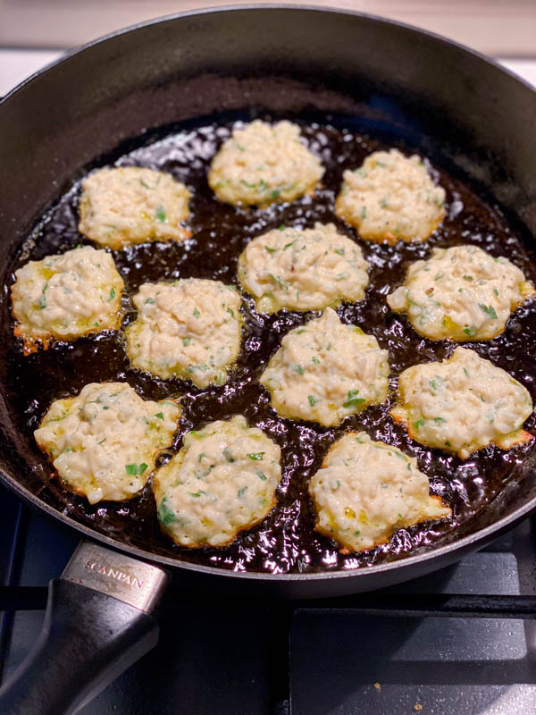 There are 12 fritters cooking in a frying pan. They are nicely golden on one side and ready to be flipped over.