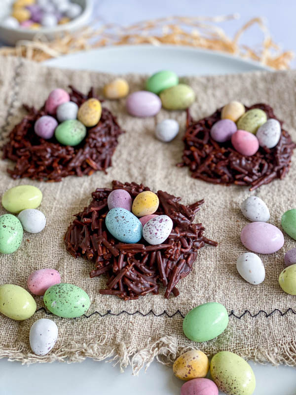 The Easter Chocolate Nests are sitting on a plate that is lined with a napkin. Scattered around the nests are mini easter eggs.
