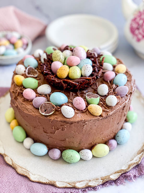 The Easter themed Chocolate Cake is now decorated with a chocolate nest in the centre which is filled with more mini easter eggs. There are also mini easter eggs placed around the base of the cake. In the background are cake plates.