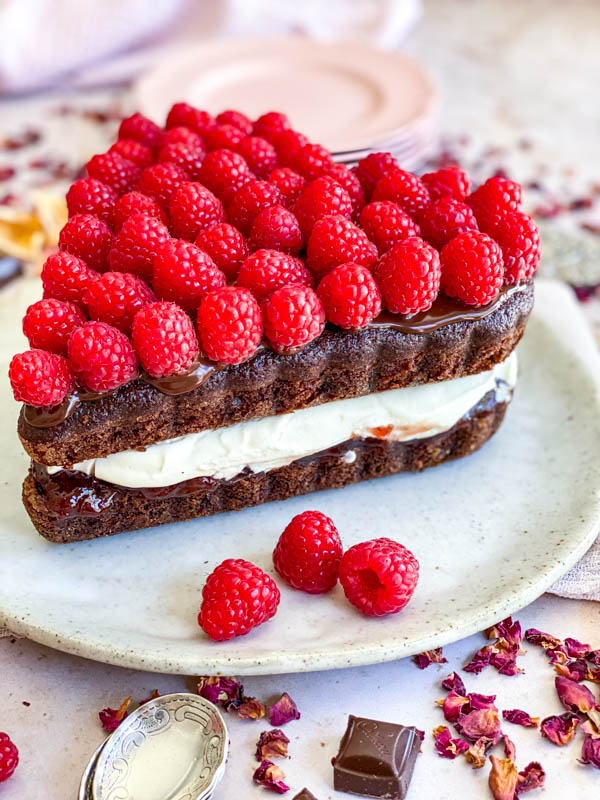 The chocolate cake is filled with jam and whipped cream and topped with a layer of ganache and fresh raspberries.