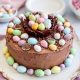 The Easter themed chocolate cake is on a plate on a table with plates and a bowl of mini easter eggs in the background.
