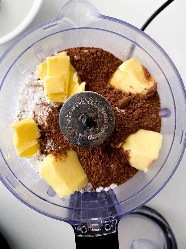 The ingredients for the chocolate icing (softened butter, icing sugar and cocoa powder) are in the bowl of a mini food processor.