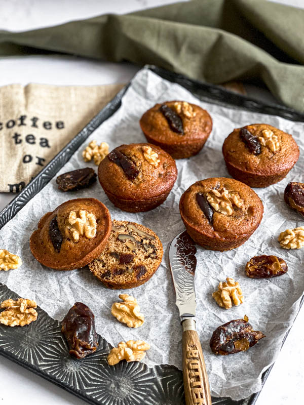 There are four whole muffins on a decorative tray with a fifth one cut open to show the inside. There is also a butter knife and a few dates and walnuts are scattered around the muffins.