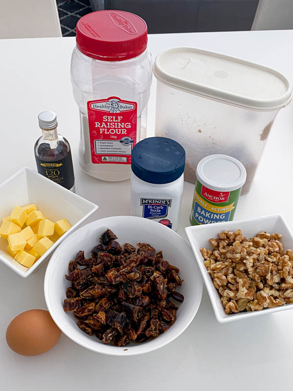 All the ingredients to make this recipe is shown here on a kitchen bench. There is self raising flour, brown sugar, chopped butter, chopped dates and walnuts, an egg, vanilla extract, baking soda and baking powder.