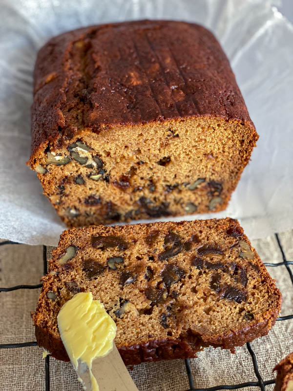 The loaf is sliced showing the inside of the loaf which is moist and packed with dates and walnuts. There is a knife with butter resting on the sliced loaf.