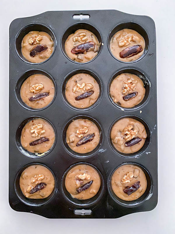 The batter is now in the greased muffin tray and each muffin is topped with a walnut and a slice of date.