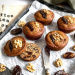 A few muffins are on a tray with one of the muffins sliced open to show the inside is moist and full of dates and walnuts. There is also a decorative butter knife with a wooden handle in the photo.