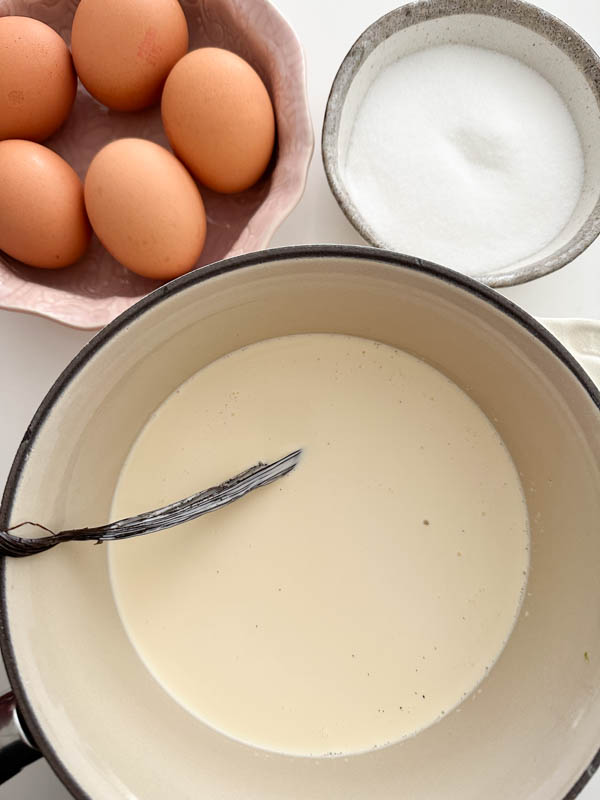 Looking down onto the ingredients required to make the ice cream - cream, milk and vanilla bean in a pot and eggs and sugar in bowls alongside.