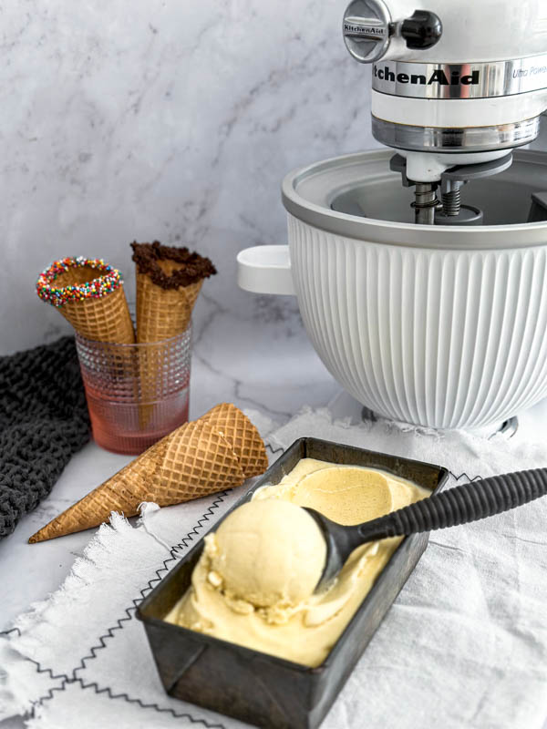The ice cream is in a tin with a scoop of ice cream on top. In the background are ice cream cones and the KitchenAid appliance.